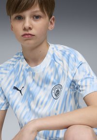 Gaiši zila un balta svītrainā sporta krekla ar apaļu kaklu, kuram ir melns Puma logo un Mančestras City emblema uz krūtīm.