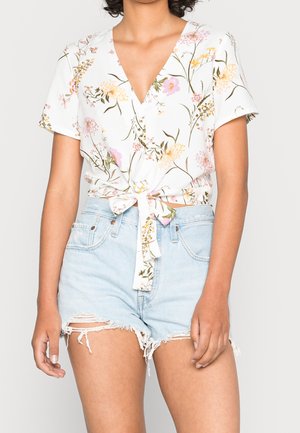 Kvinde iført en hvid kortærmet blomstret bluse bundet i taljen og lyseblå slidte denimshorts.