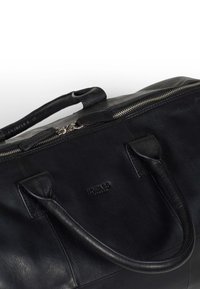 Svart läder väska med blixtlås på toppen, två hållbara handtag och präglad logotyp. Har en slät yta och enkel design.