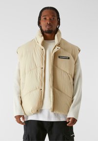 Beige puffer vest with a high collar, featuring a black logo patch, snap buttons, and diagonal quilted design. Worn over a light sweater.