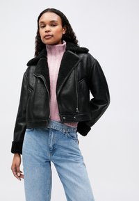 Γυναίκα που φοράει ένα μαύρο δερμάτινο μπουφάν από γούνα, ροζ πλεχτό πουλόβερ με ψηλό γιακά και ανοιχτό μπλε ψηλόμεσα τζιν μπροστά σε λευκό φόντο.