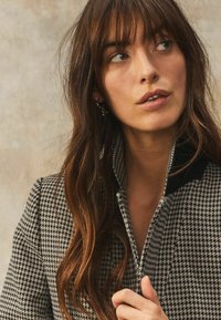 Jachetă cu model houndstooth alb-negru, cu guler înalt, realizată dintr-un material texturat și evidențiată prin accente negre contrastante.