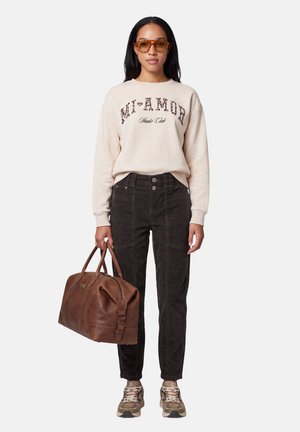 Femme portant un sweat-shirt beige avec texte, pantalon foncé, lunettes de soleil marron, baskets, tenant un grand sac de voyage en cuir marron.