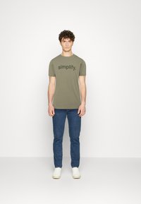 Olīvu zaļš kokvilnas T-krekls ar vārdu "simplify." treknā melnā tekstā, apvienots ar ziliem džinsiem un baltām sporta apaviem. Vienkāršs, ikdienišķs dizains.