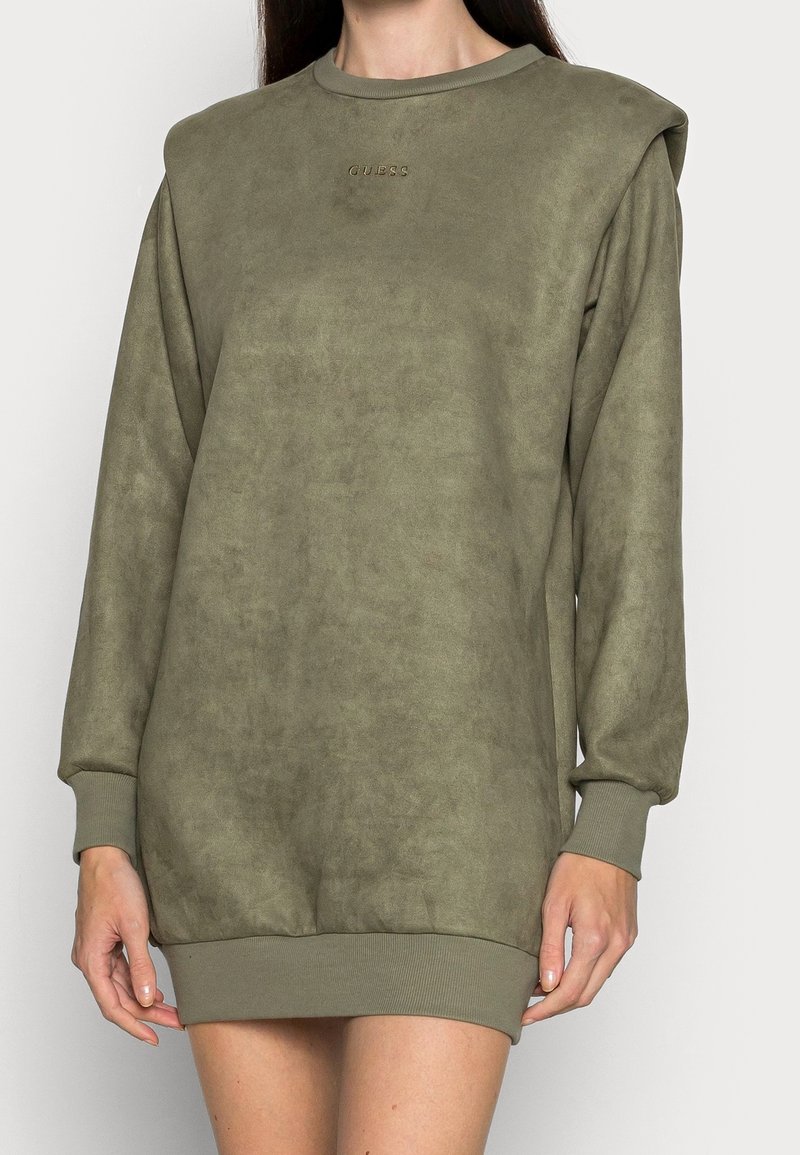Femme portant une robe sweat-shirt oversize verte à manches longues, avec épaulettes et poignets côtelés, avec un petit logo GUESS embossé sur la poitrine.