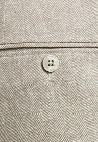 Herringbone-kuvioinen kangas vaaleanbeigellä sävyllä, jossa on tekstuuri, ja saumassa on ommeltu pyöreä, nelireikäinen valkoinen nappi.