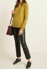 Maglione a collo alto di colore verde oliva, con bottoni decorativi sulla spalla; abbinato a pantaloni in pelle nera e sneaker nere.