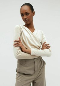 Långärmad krämfärgad topp med en omslagdesign och slät textur. I kombination med hög midja, skräddarsydda beige byxor med sidofickor.