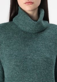 Dark green, textured knit turtleneck sweater with a wide funnel neck, showcasing a soft, warm fabric and a subtle pattern throughout.