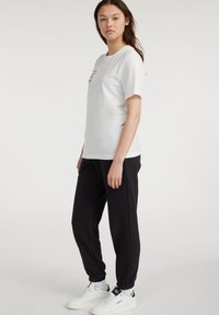 Hvit bomulls t-skjorte med korte ermer og svart trykt tekst, kombinert med svarte joggebukser med elastiske mansjetter og hvite joggesko.