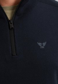 Mornarica zip-up dukserica izrađena od pletiva. Ima visoki ovratnik, crni patentni zatvarač i mali izvezeni logo na prsima.