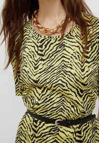 Gelbe Bluse mit schwarzen Tigerstreifen, ausgestattet mit einem runden Ausschnitt und gerafftem Stoff. Dazu wird ein schwarzer Gürtel und eine auffällige goldene Halskette getragen.