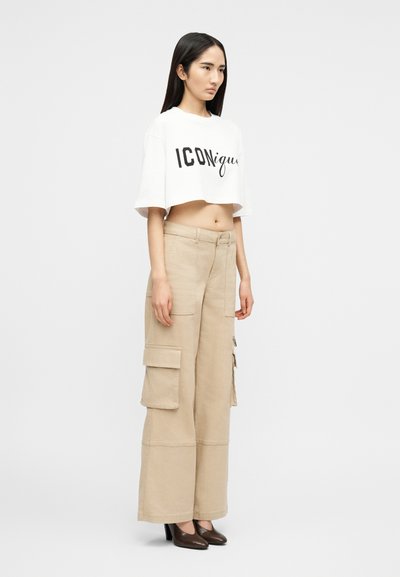 Γυναίκα με μακριά σκοτεινά μαλλιά φοράει λευκό κοντό T-shirt με το κείμενο "ICONique", μπεζ φαρδιά παντελόνια cargo και σκούρα παπούτσια με ψηλό τακούνι.