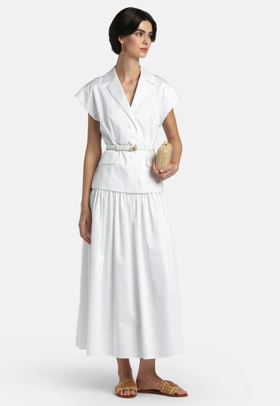 Femeie purtând un blazer cu mâneci scurte, alb, cu curea, și o fustă lungă, ținând o poșetă bej din materiale împletite, cu sandale tip papuc asortate, din același material împletit.