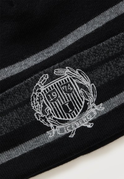 Tissu tricoté noir et gris avec un blason blanc brodé montrant "1974," feuilles et le mot "Cerberus" sur une bannière.
