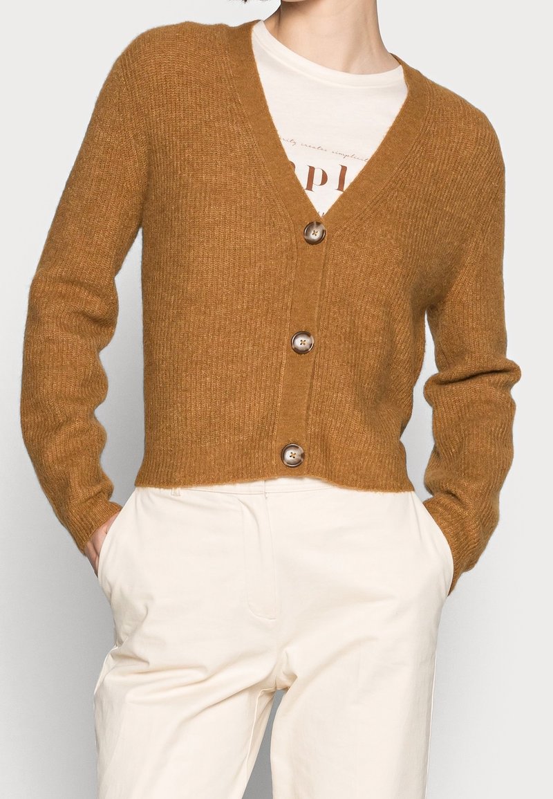 Brauner, gestrickter Cardigan mit V-Ausschnitt, fünf Knöpfen und gerippter Textur. Kombiniert mit cremefarbener Hose.