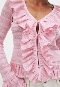 Rosa ribbad tröja gjord av strukturerad tyg, med en främre ruffle och knappstängning samt utsvängda ärmar för extra dimension.