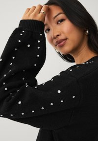 Sort strikket sweater med lange ærmer, prydet med sølvfarvede dekorative nitter på ærmerne. Blød tekstur med ribbede manchetter.