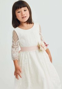Vestito bianco in pizzo con maniche trasparenti a tre quarti, una cintura rosa in vita e un accento floreale sul lato, caratterizzato da una gonna ampia con dettagli ricamati.