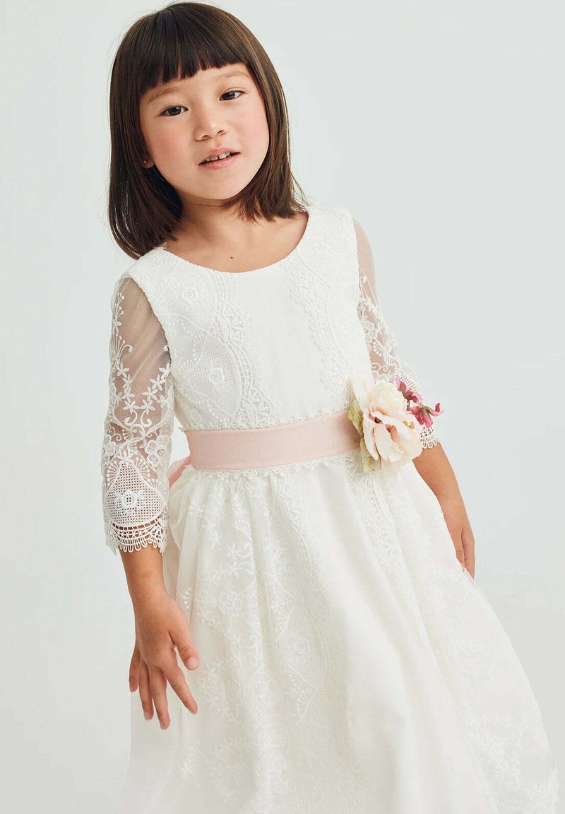 Vestito bianco in pizzo con maniche trasparenti a tre quarti, una cintura rosa in vita e un accento floreale sul lato, caratterizzato da una gonna ampia con dettagli ricamati.