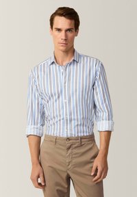 Camicia a righe blu e bianca, realizzata in cotone. Presenta maniche arrotolabili ed è abbinata a pantaloni beige chiaro.