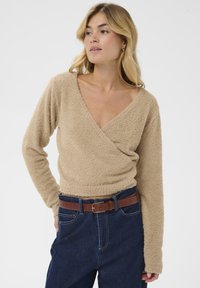 Bežna puhasta jopica s dolgimi rokavi, globokim V-izrezom in zbranim robom, v kombinaciji s temno modrimi hlačami z visokim pasom in rjavim pasom.