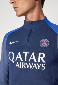 Navy athletic top with high collar and half-zip, features textured fabric, blue shoulder accents, and white lettering for "QATAR AIRWAYS" and logo.