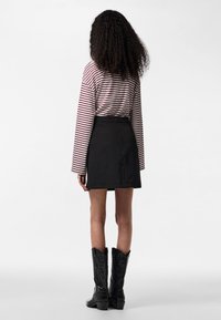 Femme aux cheveux bouclés, vue de dos, portant un haut à manches longues rouge et blanc à rayures, une jupe noire et des bottes de cowboy noires.
