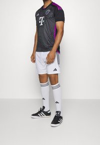 Svart fotbollströja med lila detaljer, geometriska mönster och vit logotyp, kombinerad med vita shorts och knähöga vita strumpor med ränder.