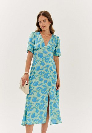 Kobieta ubrana w niebiesko-zieloną kwiecistą sukienkę midi z bufiastymi rękawami i rozcięciem z przodu, trzyma małą beżową torebkę na jednolitym tle.