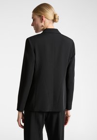 Blazer nero con un fit su misura, spalle strutturate e un tessuto liscio. Sezioni verticali evidenti e rever eleganti.