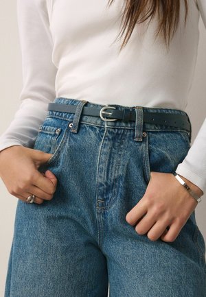 Frau trägt hoch taillierte blaue Jeans mit schwarzem Gürtel, weißes langärmeliges Oberteil, silberner Ring und Armband, Hände in der Nähe der Taschen.