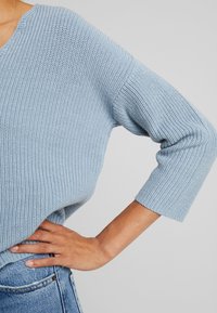Personne portant un pull en tricot bleu clair à manches trois-quarts et un jean en denim bleu, main posée sur la hanche sur un fond gris.