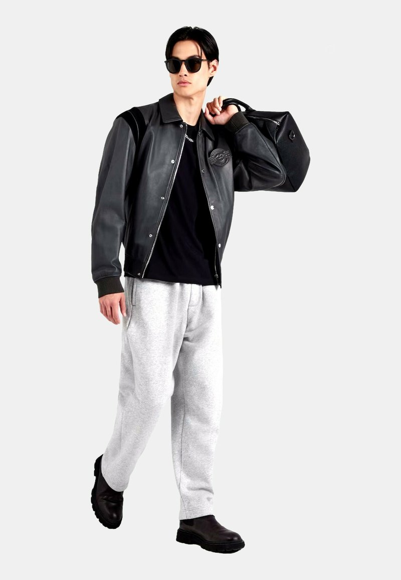 Giacca bomber in pelle nera con polsini a costine, abbinata a pantaloni della tuta grigi e stivali neri robusti. Modello con occhiali da sole, che tiene una borsa nera.