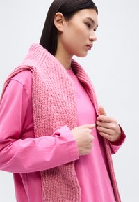 Νεαρό άτομο φορόντας ροζ μακρυμάνικο πουκάμισο και ροζ πλεκτό κασκόλ, κοιτάζοντας πλάγια ενώ τα χέρια του προσαρμόζουν το κασκόλ.