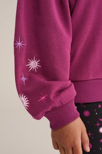 Fuksijas krāsas sporta krekls ar izšūtām debeszvaigžņu ilustrācijām uz piedurknēm, ribotām aprocēm, un kontrastējošas melnas bikses ar līdzīgiem rakstiem.