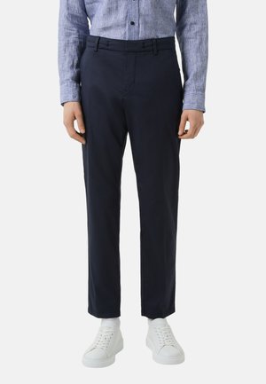 Marineblaue, maßgeschneiderte Hosen mit flacher Vorderseite und Gürtelschlaufen, aus leichtem Stoff gefertigt. Kombiniert mit weißen Sneakers für einen Kontrast.