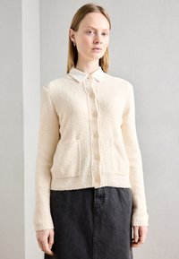 Krämfärgad cardigan i strukturerad tyg med knäppning framtill, två framfickor och en vit skjorta med krage under.