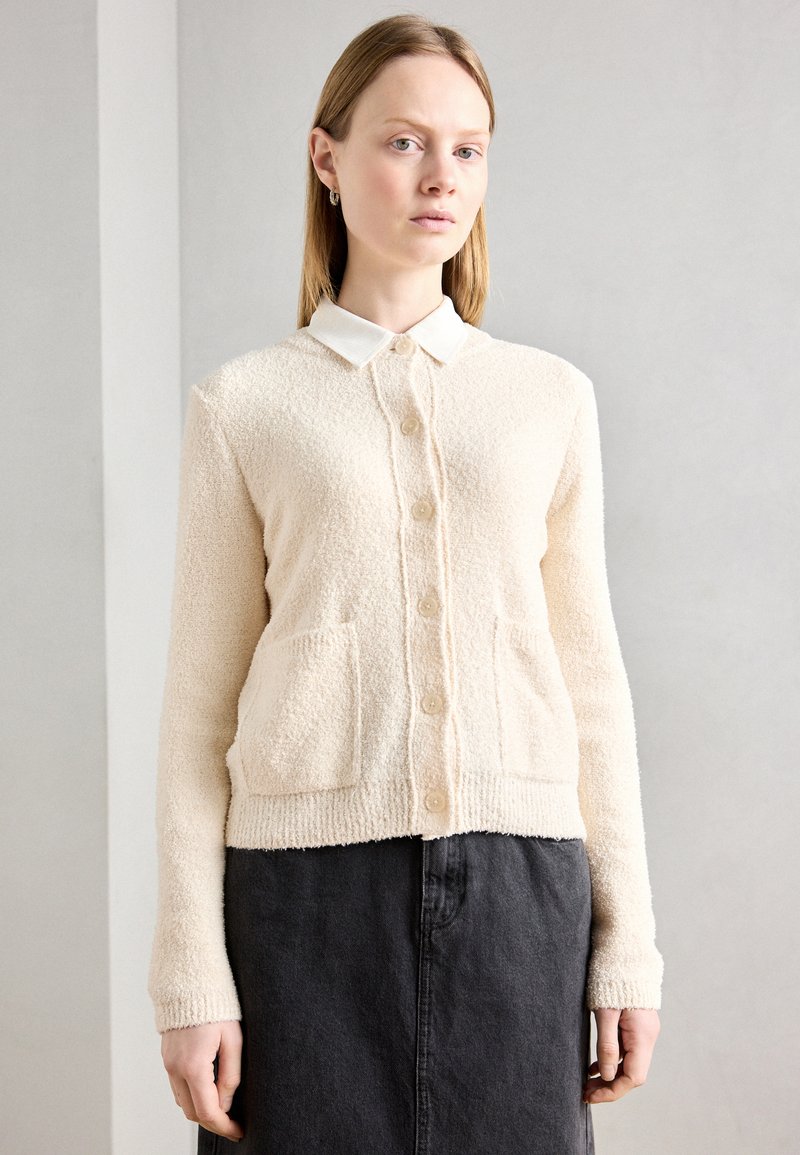 Einen cremefarbenen Cardigan aus strukturiertem Stoff mit einer Knopfleiste, zwei vorderen Taschen und einem weißen Kragenhemd darunter.