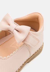 Zapato de cuero rosa con punta redondeada, borde festoneado y un lazo en la correa. Presenta una suela de goma marrón clara y un suave forro interior.