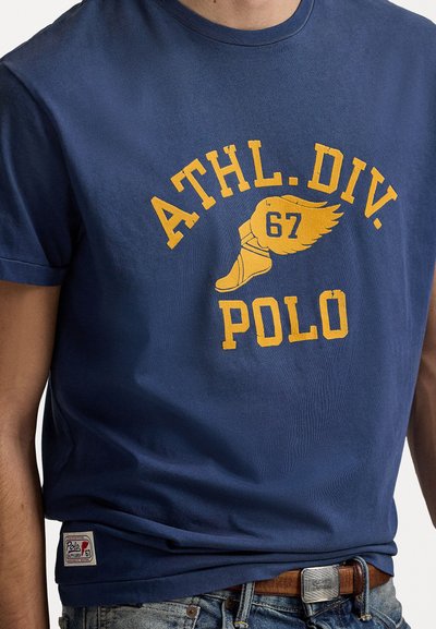 Tamsiai mėlyna medvilninė marškinė su trumpomis rankovėmis; turi geltoną užrašą "ATHL. DIV. 67 POLO" ir sparnuoto batelio grafiką. Etiketėje matomas prekės ženklo logo.