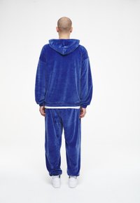 Sudadera de terciopelo azul con capucha y cordones, combinada con pantalones a juego. Ambos tienen una textura suave y un ajuste relajado. Zapatillas blancas completan el conjunto.