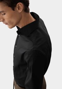 Camicia nera con bottoni, colletto alto, vestibilità slim e texture liscia. La camicia presenta cuciture sottili ed è abbinata a pantaloni marroni.