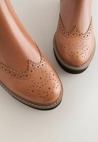 Bruine leren schoenen met geperforeerde details en gegolfde randen, voorzien van een ronde neus en contrasterende witte stiksels op de zool.