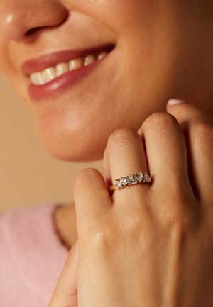 Smiling person holding hand near face, wearing gold ring with five heart-shaped clear gemstones on middle finger.