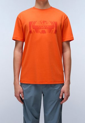 Orangefarbenes Baumwoll-T-Shirt mit kurzen Ärmeln und einem Grafikdruck in einem dunkleren Farbton. Kombiniert mit hellgrauen, locker sitzenden Hosen.