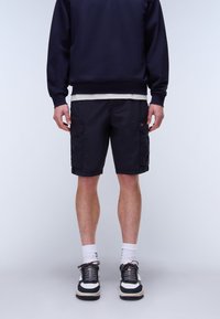 Man draagt zwarte cargobroek, witte sokken, zwart-witte sneakers en een donker sweat-shirt, staand tegen een effen achtergrond.