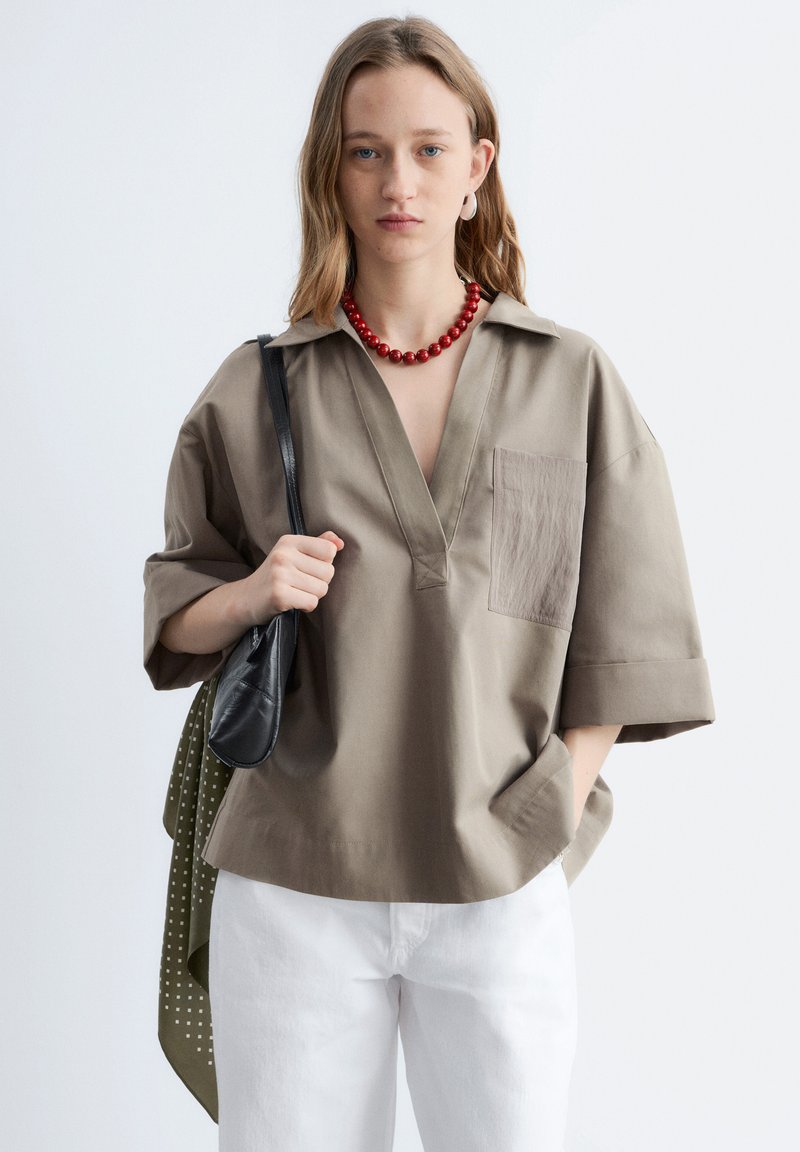Jeune femme portant une chemise ample couleur fauve, pantalon blanc, collier de perles rouges, tenant un sac noir avec une écharpe verte posée sur son épaule.