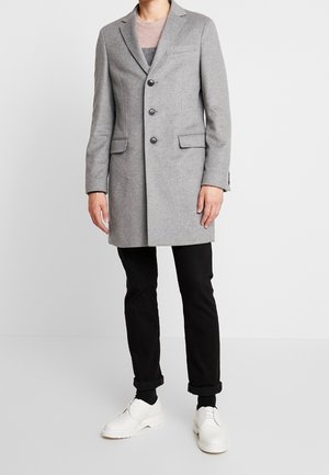 Persona che indossa un lungo cappotto grigio con bottoni, pantaloni neri risvoltati alle caviglie, calzini neri e scarpe bianche con lacci, in piedi su uno sfondo semplice.