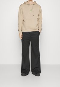 Beżowa bluza z kapturem z przednią kieszenią i sznurkami, z zielonym logo. Do tego czarne spodnie o szerokich nogawkach i czarne buty.